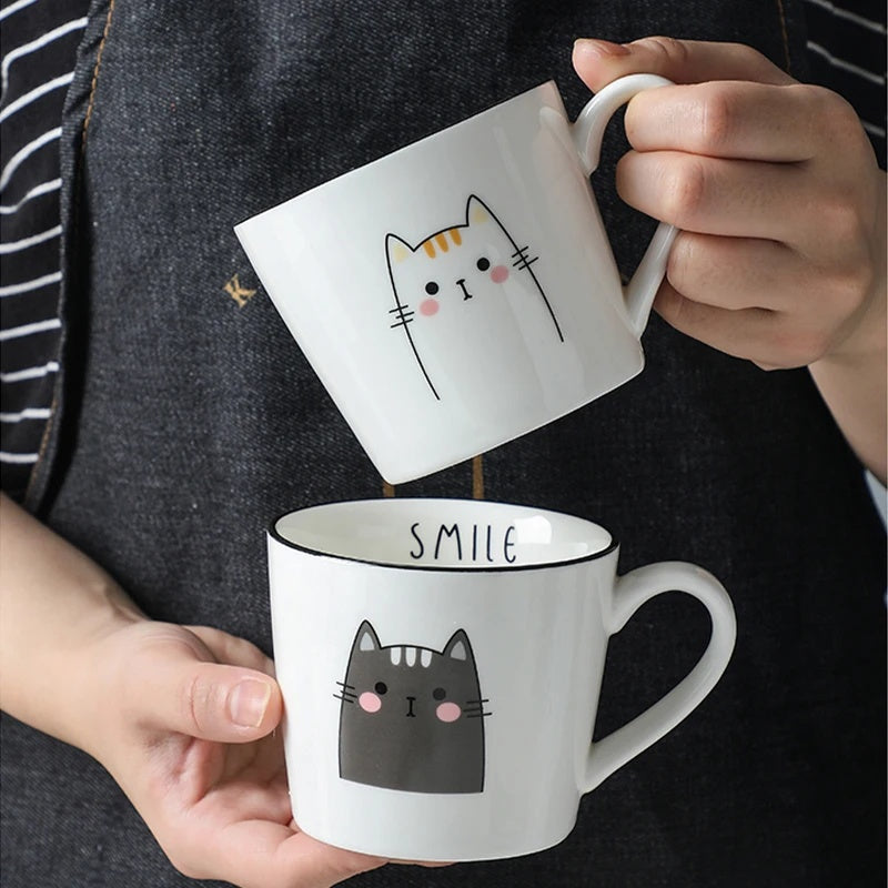 Two white mugs with cat illustrations held by a person against a dark background.